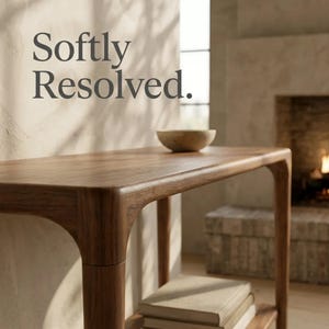 May include: A brown wooden console table with rounded edges and a lower shelf holding a stack of books. A small bowl sits on the tabletop. The text Softly Resolved. is visible in the upper left corner of the image, set against a light wall.