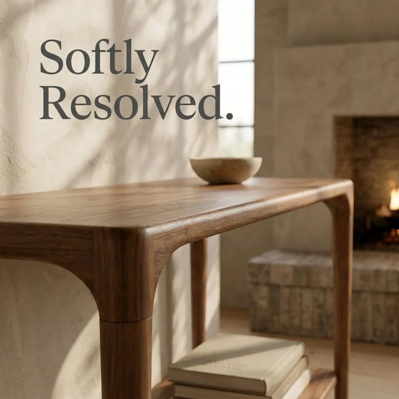 May include: A brown wooden console table with rounded edges and a lower shelf holding a stack of books. A small bowl sits on the tabletop. The text Softly Resolved. is visible in the upper left corner of the image, set against a light wall.