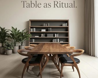 May include: A wooden dining table with a natural edge, surrounded by six curved wooden chairs with black seats. Behind the table is a tall wooden bookshelf filled with books. The wall features the text Table as Ritual in dark grey lettering.