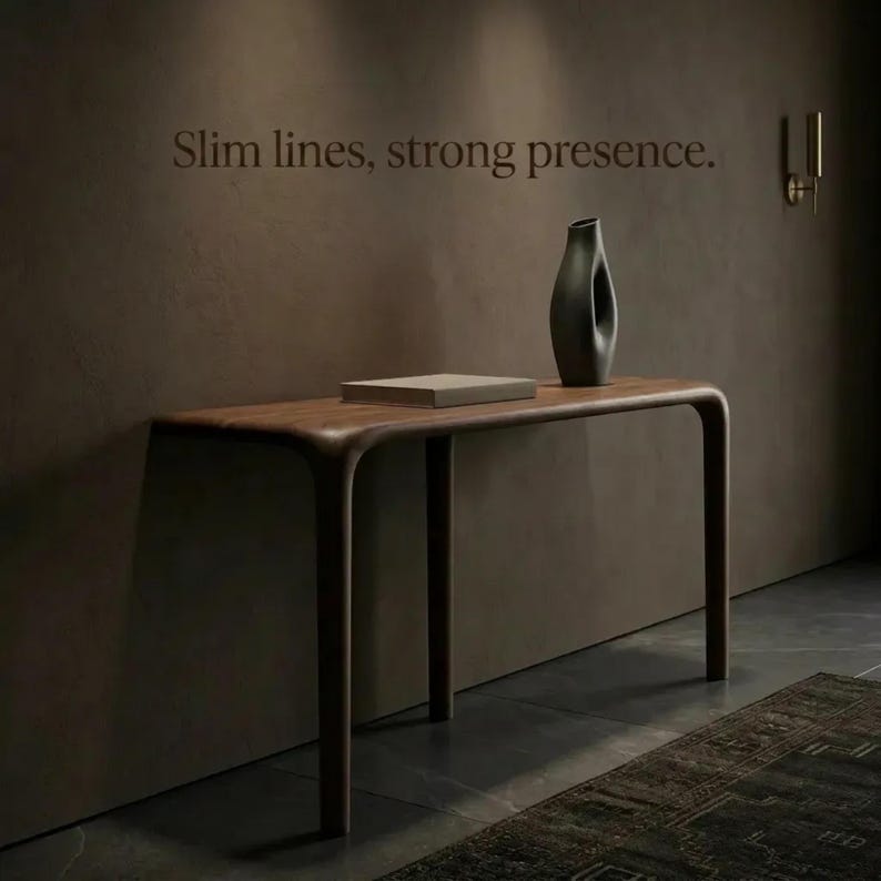 Peut inclure: Une console en bois fonc&eacute; aux bords arrondis avec quatre pieds, portant un livre rectangulaire et un grand vase sombre de forme organique. Le mur au-dessus affiche le texte Slim lines, strong presence. Une applique murale est visible.