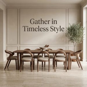 May include: A rectangular wooden dining table with eight chairs featuring curved backrests and light-coloured upholstered seats. The wall behind the table displays the dark brown text Gather in Timeless Style. A potted olive tree stands to the right.