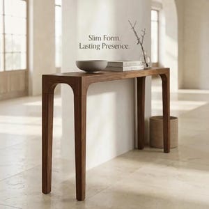Peut inclure: Une console étroite en bois avec des pieds arqués et une finition brun foncé lisse. Sur le dessus se trouvent un bol gris, deux livres empilés et un vase en verre avec une branche fine. Le texte Slim Form. Lasting Presence. est visible.