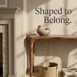 May include: A beige ceramic vase with an asymmetrical, organic shape sits on a wooden console table. Below the table is a woven basket and a stack of books. The wall features the dark grey text "Shaped to Belong." in a serif font.
