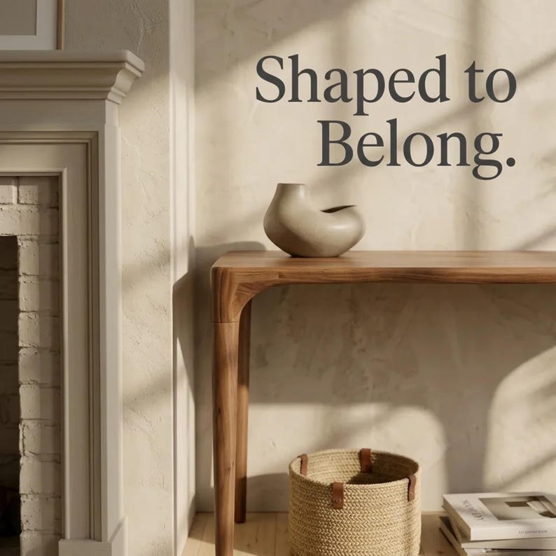 May include: A beige ceramic vase with an asymmetrical, organic shape sits on a wooden console table. Below the table is a woven basket and a stack of books. The wall features the dark grey text "Shaped to Belong." in a serif font.