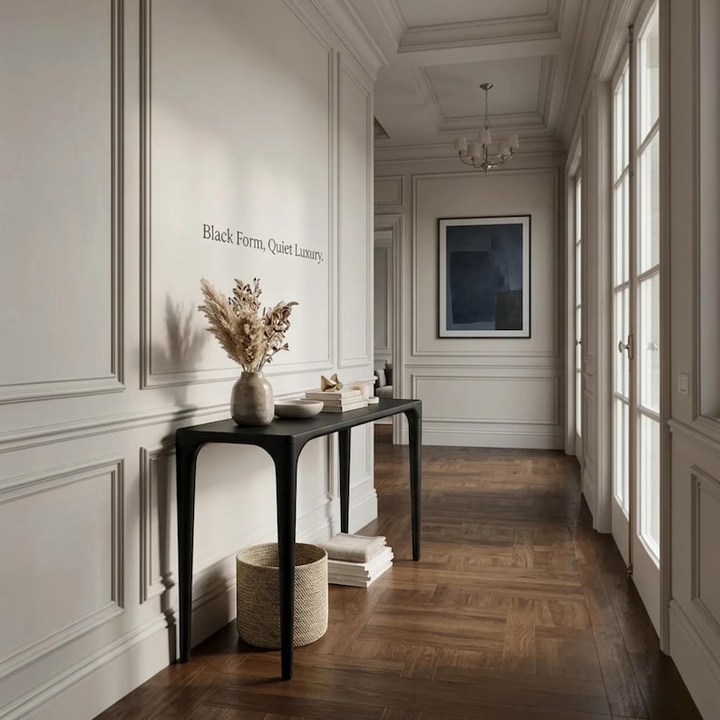 Puede incluir: Una mesa consola negra de cuatro patas delgadas se apoya contra una pared con paneles que presenta el texto Black Form, Quiet Luxury. Sobre la mesa hay un jarr&oacute;n de cer&aacute;mica con hierbas secas y una pila de libros, con una cesta tejida debajo.