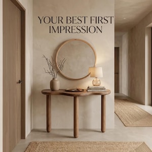 Puede incluir: Una mesa consola de madera con tres patas redondeadas, un espejo de pared redondo, un jarrón beige con ramas desnudas, una lámpara de mesa con pantalla crema y una pila de libros. La pared muestra el texto negro: YOUR BEST FIRST IMPRESSION.