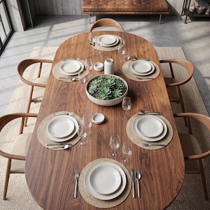 May include: An oval walnut dining table with six wooden chairs featuring curved backrests. The table is set with six woven placemats, white ceramic plates, stainless steel cutlery, clear glass stemware, and a central bowl of green succulents.