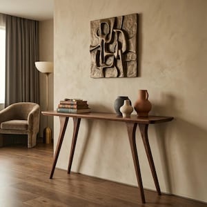 May include: A dark wood console table with four angled legs, topped with a stack of three books, a magnifying glass, and three ceramic vases in black, cream, and terracotta. A square bronze-toned wall relief with abstract raised shapes.