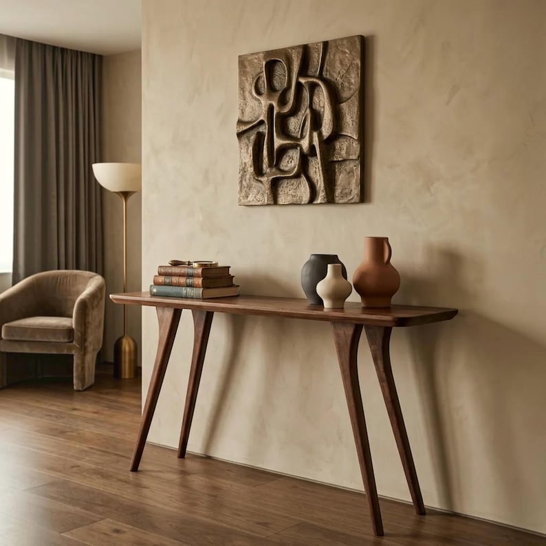May include: A dark wood console table with four angled legs, topped with a stack of three books, a magnifying glass, and three ceramic vases in black, cream, and terracotta. A square bronze-toned wall relief with abstract raised shapes.