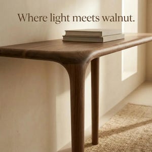 Peut inclure: Une console en bois de noyer brun fonc&eacute; avec des bords arrondis et des pieds cylindriques. Deux livres rectangulaires empil&eacute;s reposent sur la surface. Le texte "Where light meets walnut." est imprim&eacute; en lettres brunes au-dessus.
