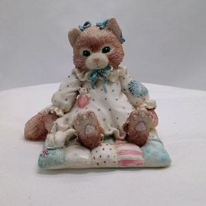 May include: A ceramic figurine of a cat wearing a blue and white polka dot dress and a blue bow. The cat is sitting on a patchwork quilt with two small stuffed mice.