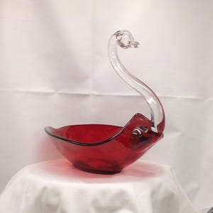 Puede incluir: Un tazón de vidrio rojo en forma de cisne con un asa de cuello de cisne de vidrio transparente.