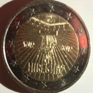 Può includere: Una moneta da 2 euro in argento e oro con la scritta "2010" e "EUROPA" sull'anello esterno. Il centro della moneta presenta un'immagine stilizzata di un edificio con un design a raggiera e la scritta "SLOVENIJA".