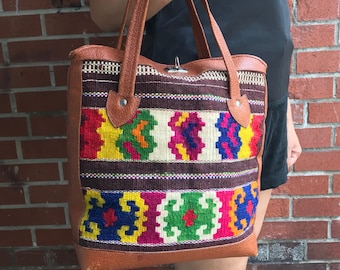 May include: A brown leather tote bag with a colourful woven fabric panel. The fabric features a geometric pattern in shades of red, green, blue, yellow, and white.