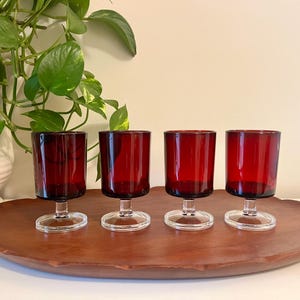 Può includere: Quattro bicchieri da vino in vetro rosso con stelo e base trasparente su un vassoio di legno.