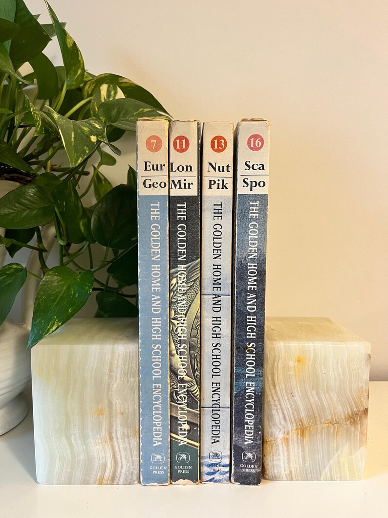 May include: Four vintage encyclopedias, volumes 7, 11, 13, and 16, are displayed upright. The books are in a row, with titles including "Eur Geo Mir" and "Nut Pik Spo." They are held in place by a light-colored, rectangular bookend. The books are from "The Golden Home and High School Encyclopedia."