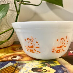 May include: A white milk glass mixing bowl featuring a repeating orange graphic print of an elephant balancing on a ball. The bowl has a smooth, rounded rim and a slightly tapered base, with a clean, glossy finish throughout the exterior surface.