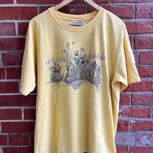 Peut inclure: Un t-shirt jaune à manches courtes avec un motif floral. Le graphisme représente une nature morte de fleurs en pots et un panier en osier. Le t-shirt est suspendu à un cintre en bois contre un mur de briques rouges.