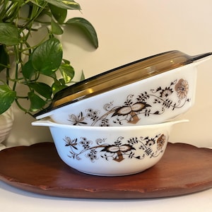 Puede incluir: Dos platos para hornear de cerámica blanca con diseños florales marrones y tapas. El plato superior tiene una tapa de vidrio marrón. Los platos están apilados en una bandeja de servir de madera.