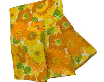 Pode incluir: Tecido floral vintage dobrado com grandes flores laranja, amarelas e castanhas e folhas verdes sobre um fundo branco. O tecido tem uma estética retro, perfeito para projetos de costura ou decoração de casa.