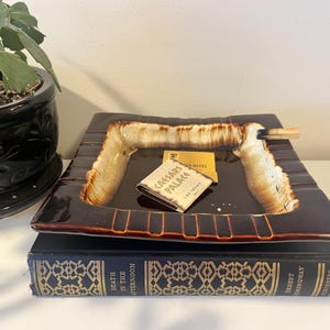 Puede incluir: Un cenicero cuadrado marrón oscuro con un borde texturizado de color crema y una pequeña ranura rectangular. Dentro hay una cerilla y una caja de cerillas del hotel Caesars Palace. El cenicero descansa sobre un libro titulado "Death in the Afternoon."