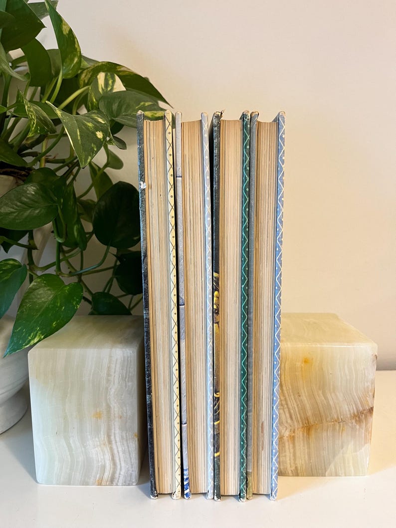 May include: A stack of vintage books with aged, yellowed pages. The spines feature decorative patterns in blue, green, and gold. The books are flanked by two square, marble bookends.