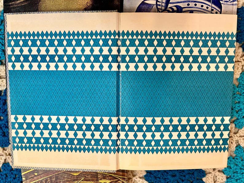 May include: Open book with a teal and cream geometric pattern. The design features rows of white diamonds and teal diamond patterns. The book is open, revealing the interior pages with the same design.