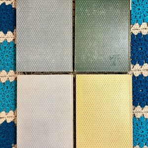 May include: Four playing card decks with diamond patterns in gray, green, off-white, and yellow. The cards are arranged in a square, with each deck's back facing up. The background is a blue and cream crocheted blanket.