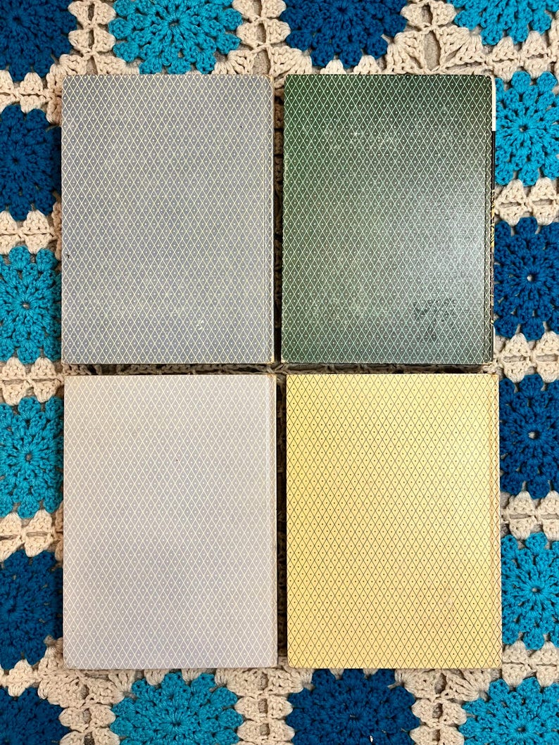 May include: Four playing card decks with diamond patterns in gray, green, off-white, and yellow. The cards are arranged in a square, with each deck's back facing up. The background is a blue and cream crocheted blanket.