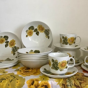 Puede incluir: Un juego de vajilla de cerámica blanca con un patrón floral con girasoles amarillos y verdes. El juego incluye cuencos, platos, tazas y platillos.