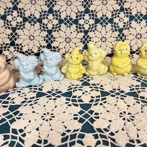 May include: A selection of ceramic figurines in different shapes and colours. The figurines include a pink pig, blue clowns, a yellow gnome, a yellow clown, and two yellow dog-like figures. They are arranged on a white lace tablecloth.