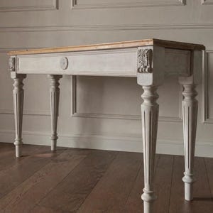 May include: A light grey console table with a natural wood top. The table has ornate carved details on the legs and corners, and a small circular design on the front. The table is approximately 152 cm long.