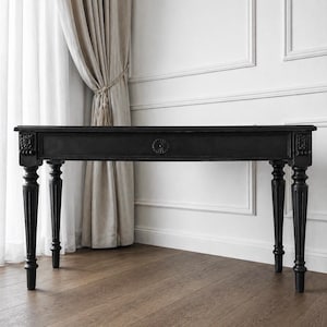 Peut inclure: Bureau en bois noir avec des détails sculptés et quatre pieds cannelés. Le bureau comporte un tiroir central avec un motif floral et des sculptures décoratives sur les coins. Le bureau est posé contre un mur blanc et un parquet.
