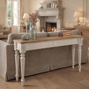 Puede incluir: Una consola blanca y de madera con detalles tallados y patas torneadas. La mesa está colocada frente a un sofá gris y está decorada con botellas de vidrio y libros. La habitación tiene una chimenea y una paleta de colores neutros.