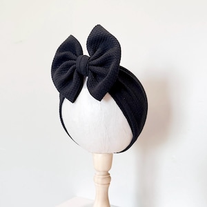 May include: A black baby headband with a large bow. The bow is textured and sits atop a matching black band. The headband is displayed on a white mannequin head with a wooden stand, against a white background.