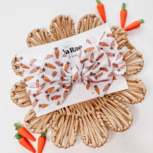 May include: A white headband with a bow, featuring a carrot print. The headband is on a woven straw background with small orange carrots around it. The text "La Rae de bows" is printed on the headband.