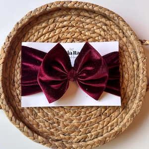 May include: A deep burgundy velvet headband featuring a large, centered bow. The headband is displayed on a white rectangular card with the text "ia Ra" visible in black print, centered on a round, woven, light brown natural fiber tray.