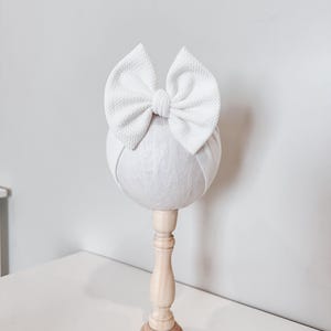 May include: A white fabric bow headband on a white mannequin head. The mannequin is on a wooden stand.