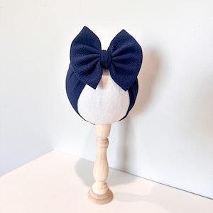 May include: A navy blue baby headband with a large bow. The bow is textured and sits atop a white, oval-shaped form. The headband is displayed on a light wooden stand against a white background.