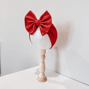 May include: A red glitter bow headband on a white mannequin head, displayed on a light wood stand. The headband features a large, sparkly bow. The background is a plain white wall and white surface.