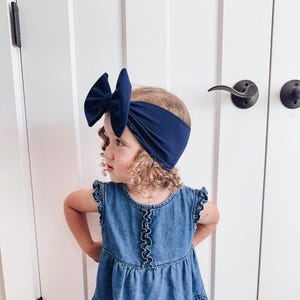 May include: A navy blue baby headband with a large bow. The headband is made of a textured fabric and is worn by a young child wearing a denim dress. The child is standing in front of a white door.
