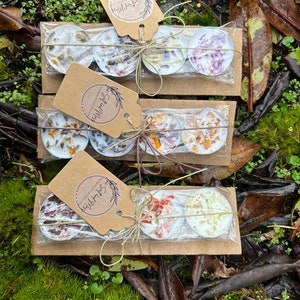 May include: Three sets of three small, round, white tea light candles with dried flowers and herbs inside. Each set is wrapped in brown paper with a twine bow and a brown paper tag that says "Wild Free & Soul".