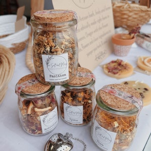 May include: Four glass jars filled with loose leaf tea blends. The jars are labeled with the names "Aphrodite", "Pasithea", "Tyche", and "Thea". The jars are labeled with the text "The Salt and Moss" and "Loose leaf tea blends".