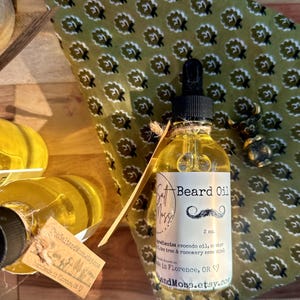 May include: A clear glass bottle of beard oil with a black dropper, labeled "Salt Moss!" and "Beard Oil." The label includes a mustache graphic and the text "2 oz." The bottle sits on a wooden surface with a green patterned fabric.