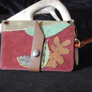 May include: A burgundy leather wallet with a flap closure and a brown leather strap. The wallet is decorated with various cutouts of leaves and a brown leather oak leaf. The wallet has a metal clasp and a brown leather strap with a metal ring.