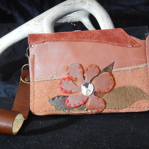 May include: A small, brown leather wallet with a flower design. The flower is made of different colored leather pieces and has a brown leather center. The wallet has a brown leather strap.