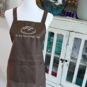 Pode incluir: Um avental castanho com um desenho bordado branco de um pão e o texto "in my sourdough era".