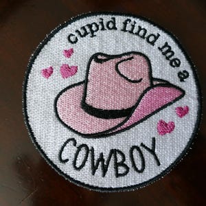 Puede incluir: Un parche circular blanco con corazones rosas y el texto "cupid find me a cowboy" en negro. Un sombrero de vaquero rosa con una banda negra está bordado en el centro.