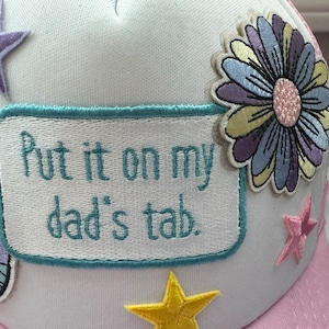 May include: White trucker hat with pink mesh back. The hat has several embroidered patches, including a rainbow, stars, a flower, and a patch that says "Put it on my dad's tab."
