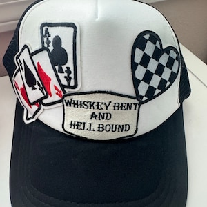 May include: A black and white trucker hat with a white mesh back. The hat has several patches, including playing cards, a checkered heart, and a patch that says "Whiskey Bent and Hell Bound".